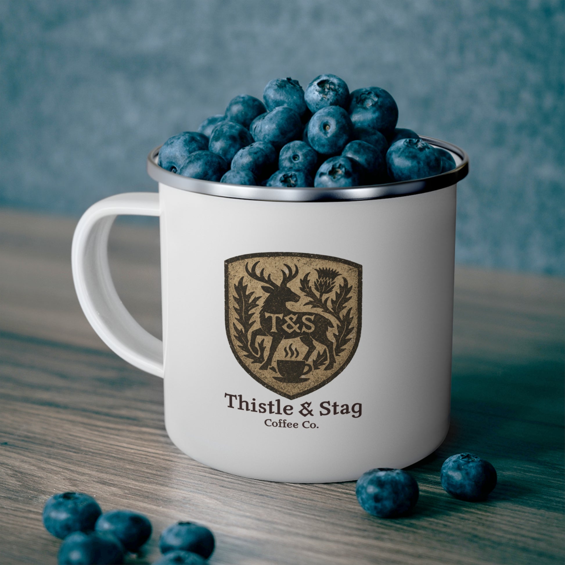 White mug with 'Thistle & Stag Coffee Co.' logo filled with blueberries on a wooden surface.